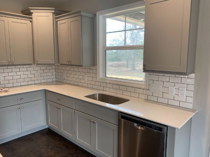 White kitchen cabinets, stainless steel sink under a window, tile backsplash, stone countertop, built-in dishwasher, drawers and cupboards