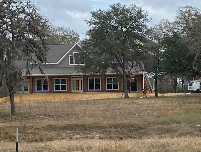 Two-story custom home with light siding, large windows, gabled roof, manicured lawn, and mature trees in the background