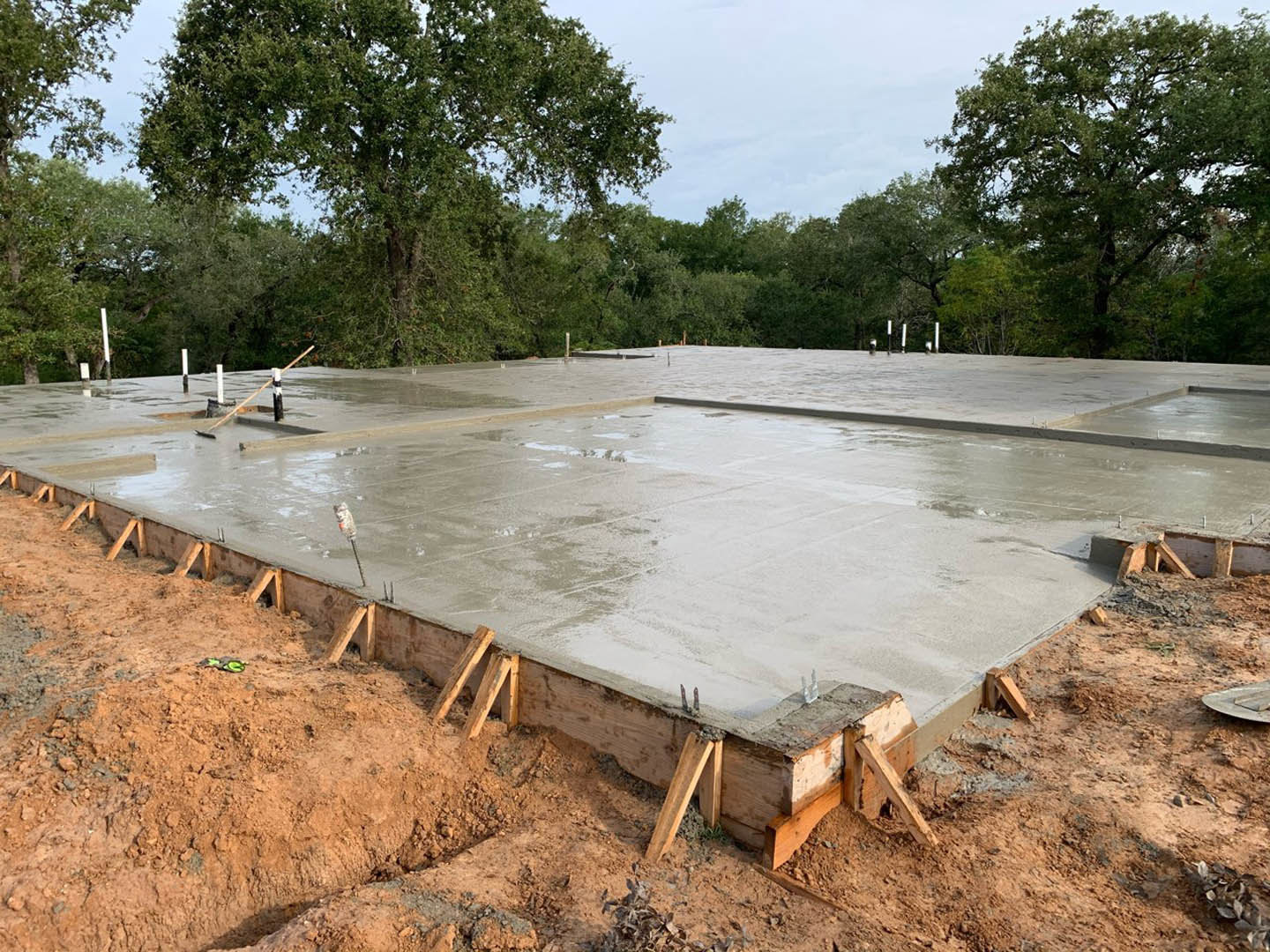 Fresh concrete slab being poured into a rectangular foundation, surrounded by dirt and construction materials, with a leafy tree visible nearby.