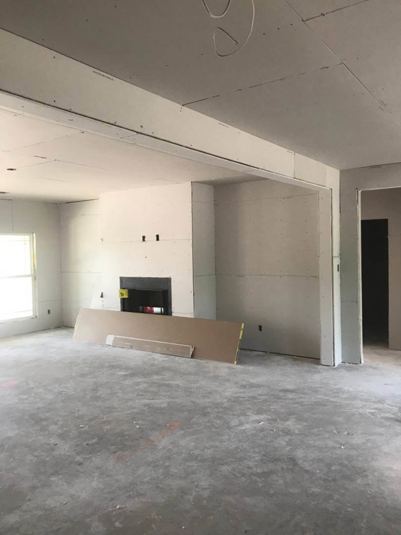 Modern living room with smooth concrete flooring, white plaster walls, built-in fireplace, large white-framed window, and minimal furnishings including a brown board and black