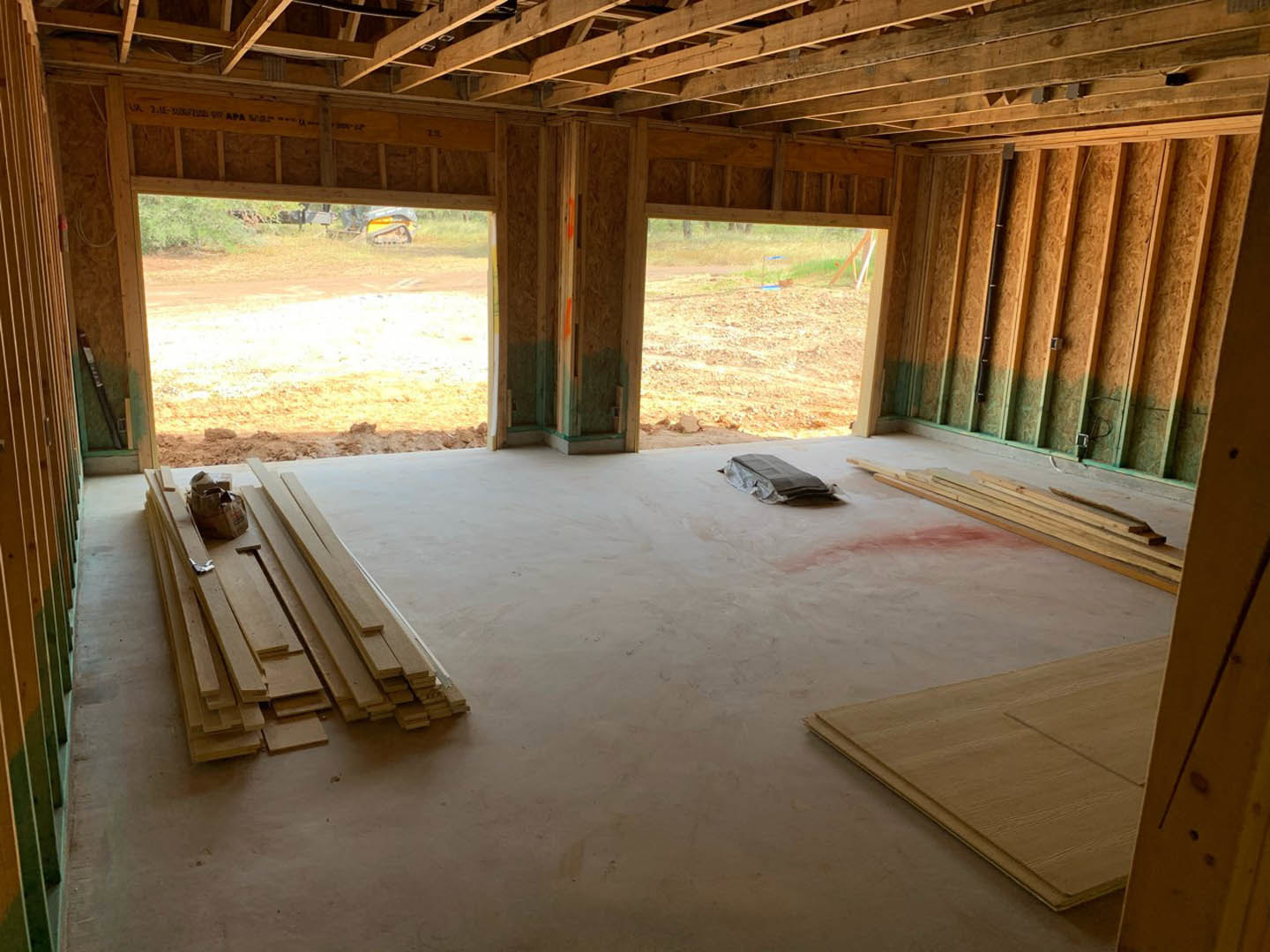 Sunlit room with unfinished wood plank flooring, scattered lumber piles, folded newspaper on a table, large windows, and exposed beams.