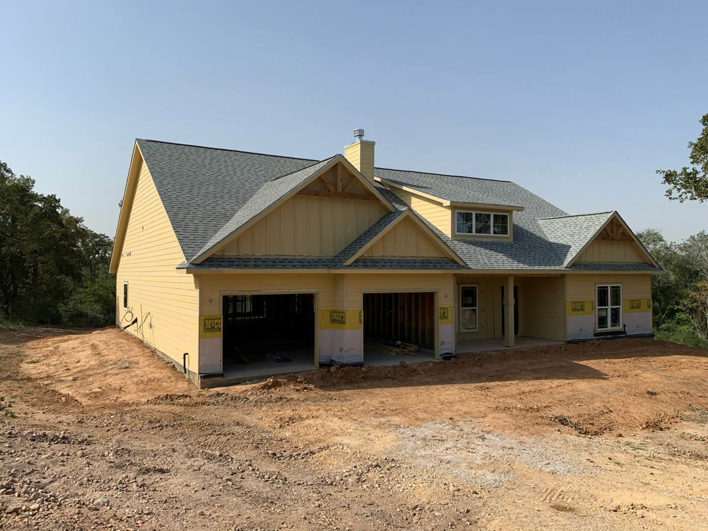 Partially built house with exposed framing, attached garage, dirt construction site, and surrounding trees in the background