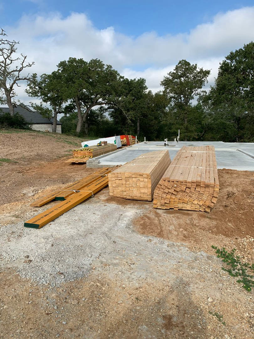 Stack of unfinished wood planks on dirt ground at residential construction site, surrounded by grass and bare tree branches under cloudy sky