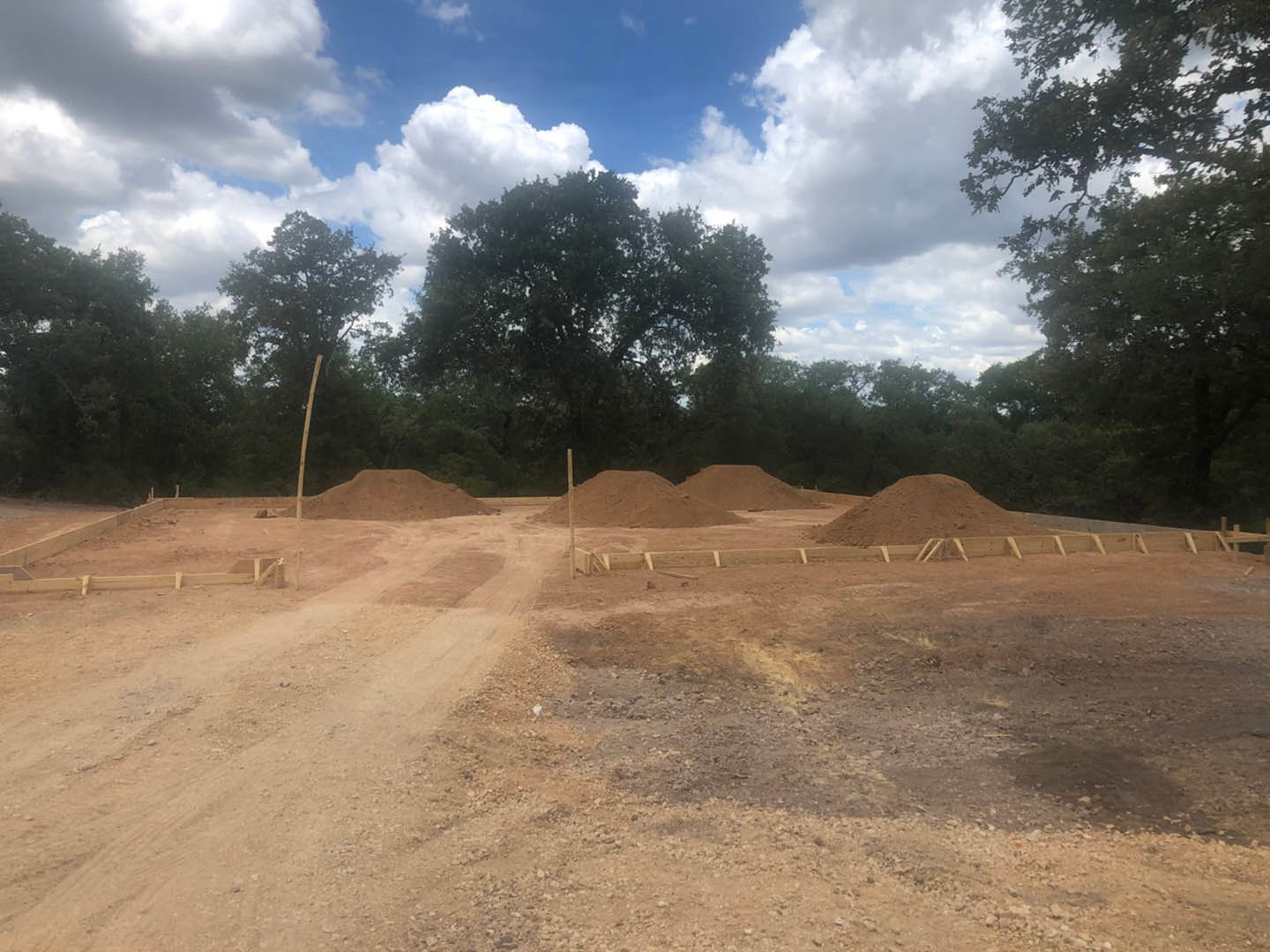 Dirt road bordered by piles of soil and leafy trees under a partly cloudy sky