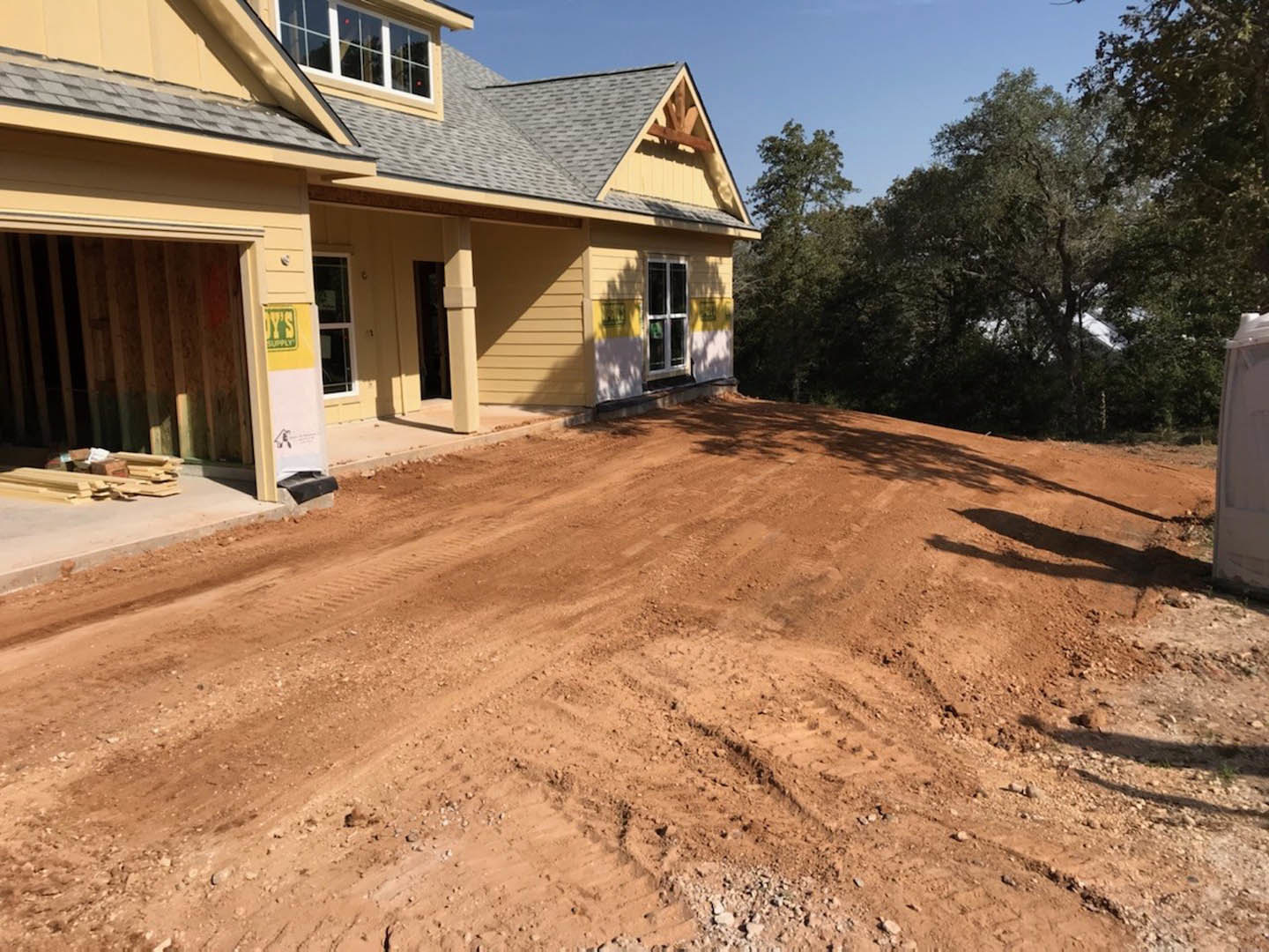 Two-story custom home with white-framed windows, surrounded by dirt landscaping and mature trees, unfinished driveway with visible tire tracks