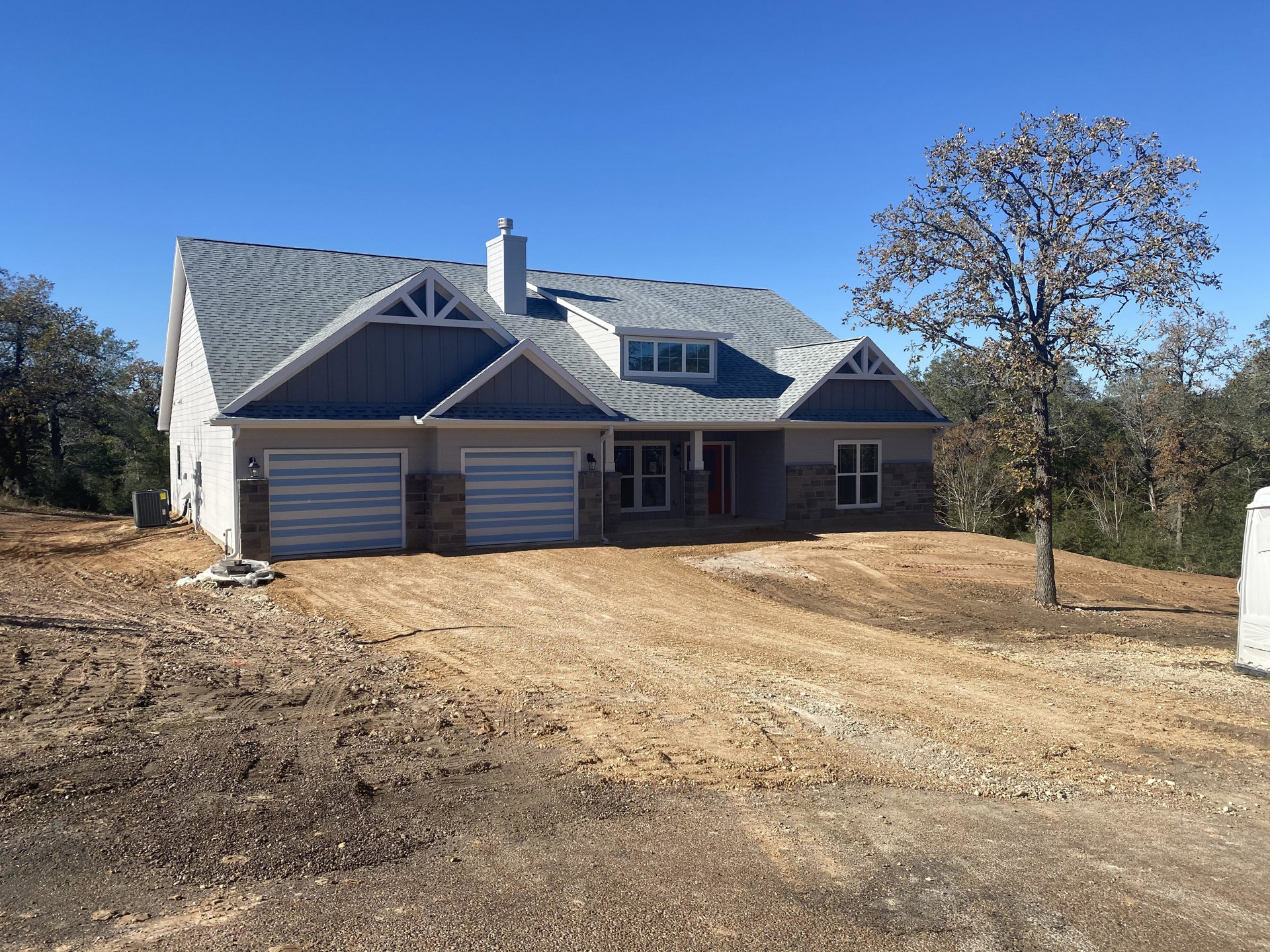 Two-story house with gray siding, white trim, and glass-paneled front door; attached garage with white doors; brick chimney on shingled roof; mature tree in background; dirt yard