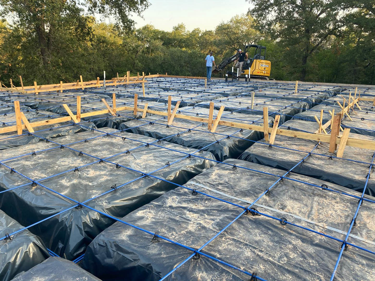 Large black tarp covering foundation area, yellow tractor parked behind, blue pipes and ropes visible, two men working outdoors near trees and construction materials.