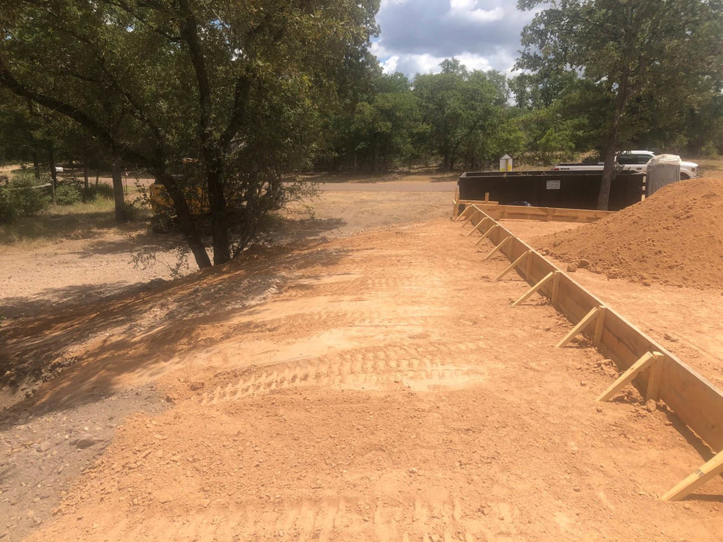 Dirt road with tire tracks, pile of soil, scattered wooden planks, and dense trees under a blue sky with clouds