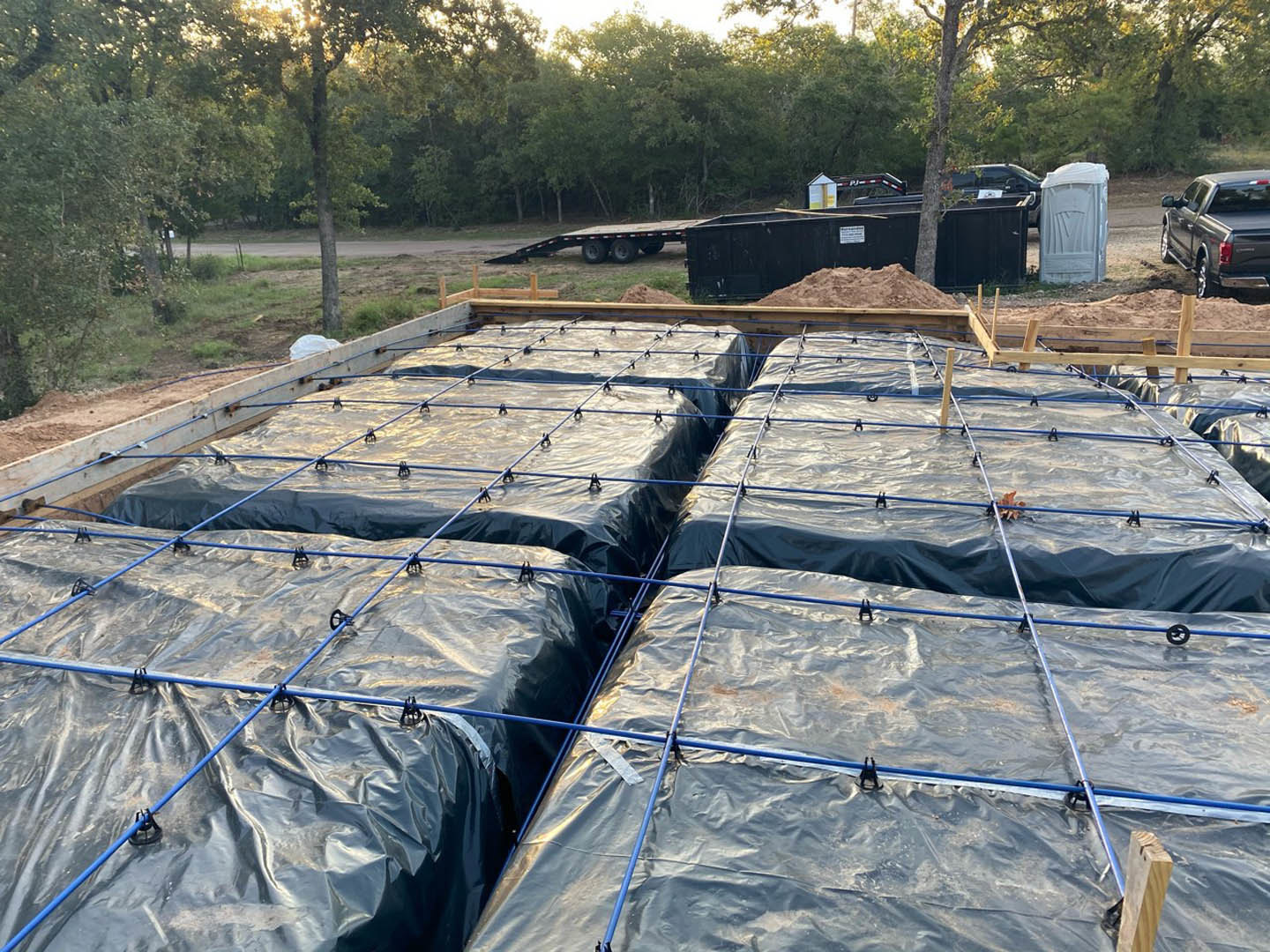 Large black plastic bags resting on a wooden frame near a pile of dirt, with a dumpster and trees in the background.