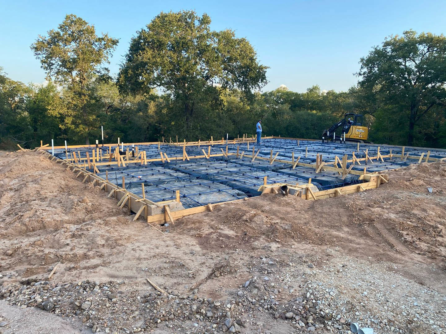 Partially built custom home with exposed foundation, piles of dirt, blue plastic coverings, and a man standing near the structure; large leafy tree in background.