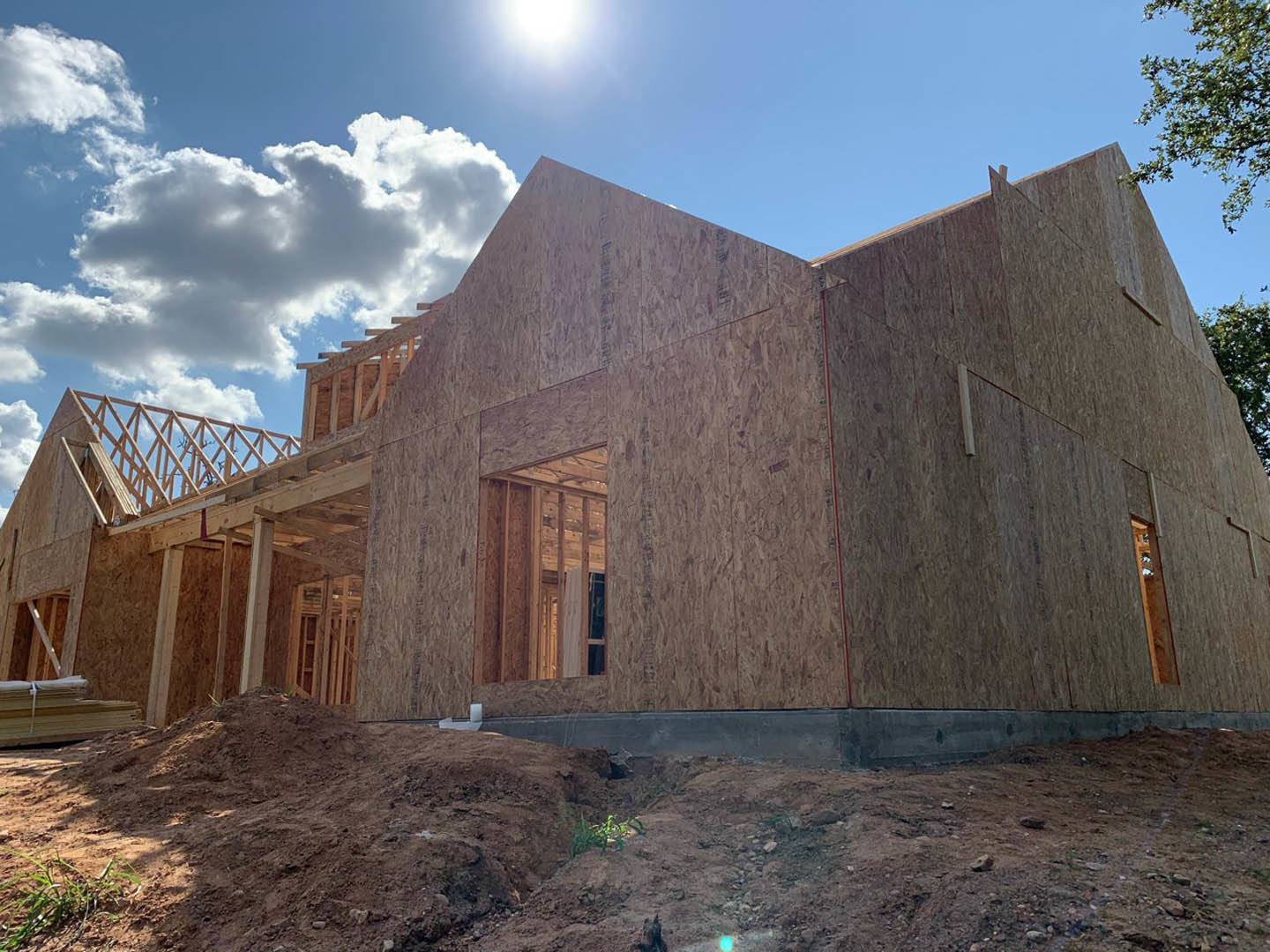 Wood-framed house under construction on a dirt hill, brick exterior partially visible, blue sky with scattered clouds, small tree and ground vegetation in foreground