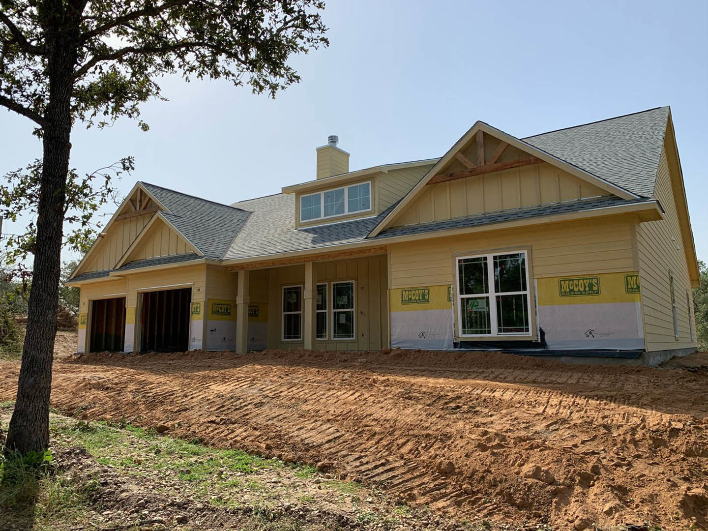 Framed house under construction with exposed wood, several installed windows, dirt yard, surrounding trees, and visible chimney structure