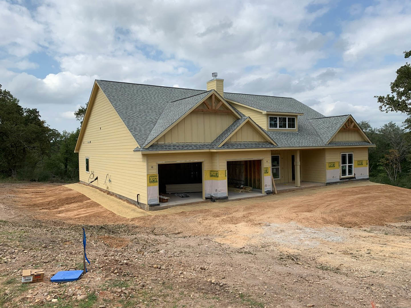 Partially built house with exposed framing, attached garages, brick chimney, multi-pane windows, and dirt yard; blue square object sits on ground near construction site.