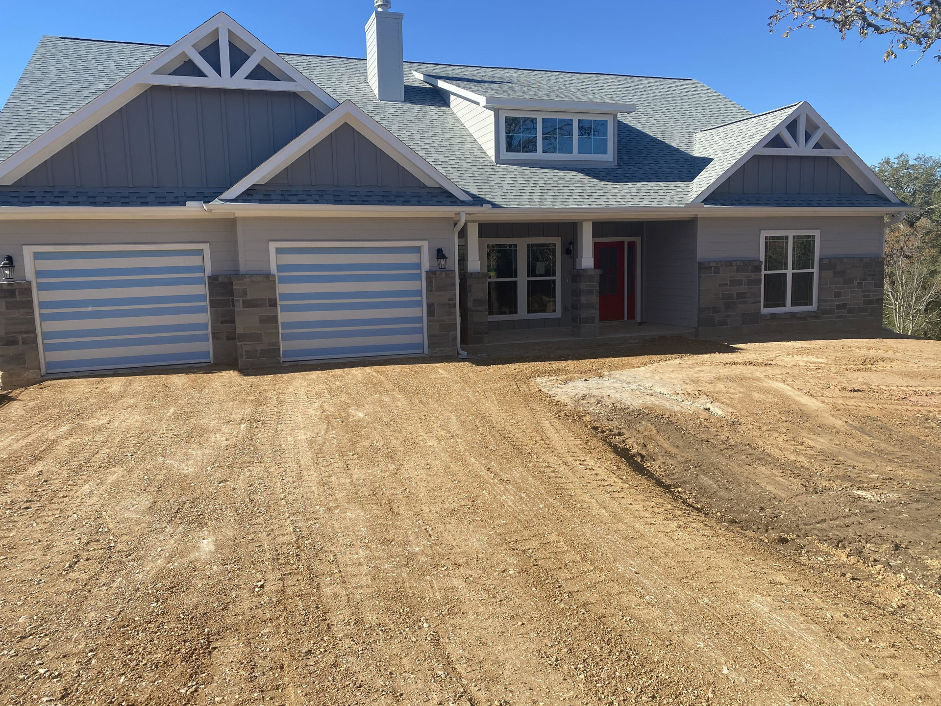 Two-story cottage with blue and white siding, red front door with glass panes, white-framed windows, attached garage, paved driveway, and mature trees in the yard