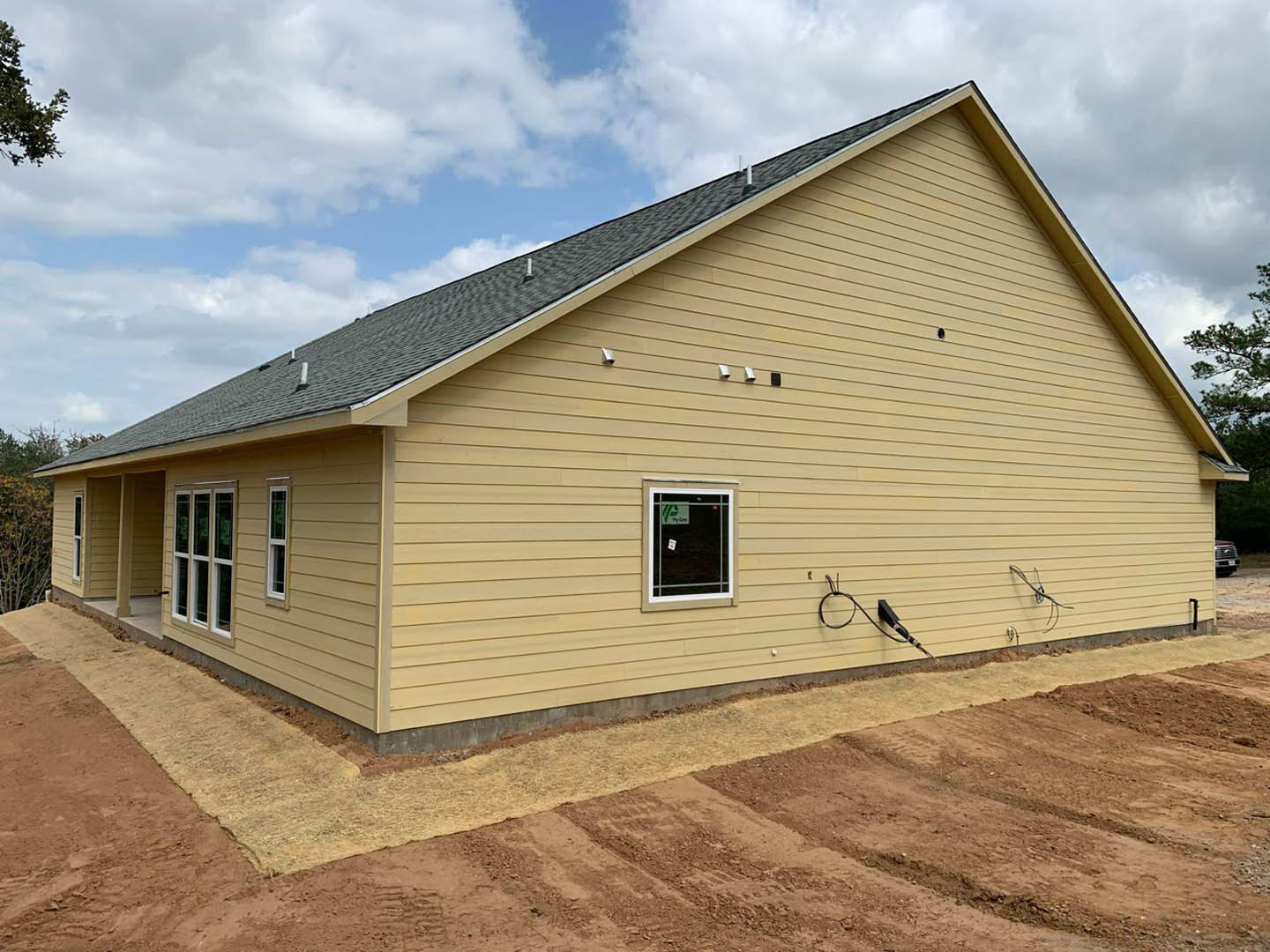 Yellow house under construction with exposed dirt patch, black cable attached to exterior wall, single window displaying a sign, blue sky and clouds overhead