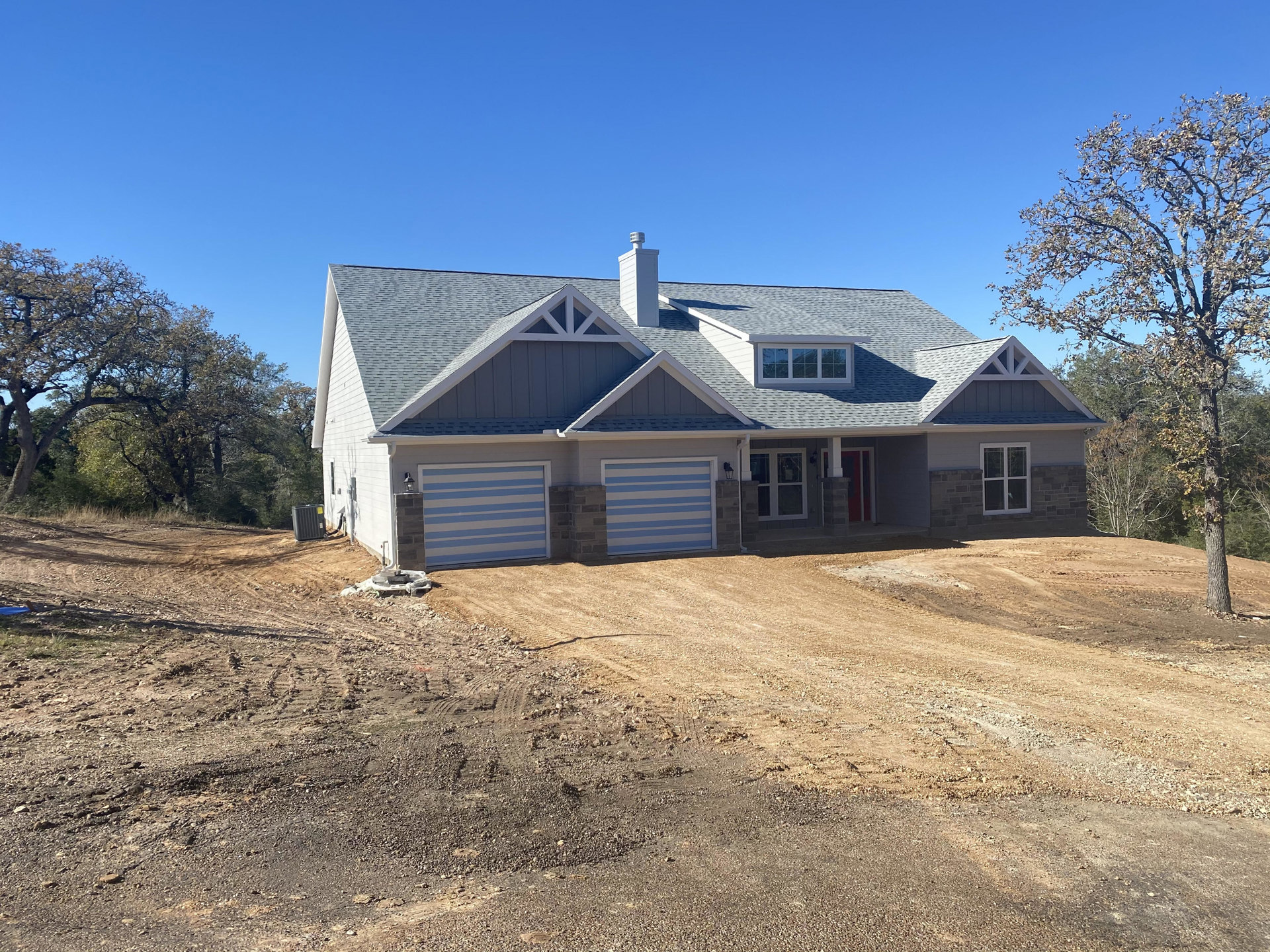 Two-story house with attached garage, white chimney, white-framed windows, gray siding, tree beside the home, dirt driveway leading to the entrance