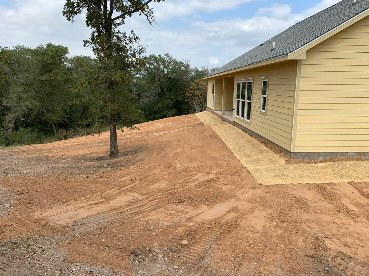 Partially built yellow house with exposed framing, dirt path leading to entrance, leafy tree nearby, blue sky and scattered clouds overhead