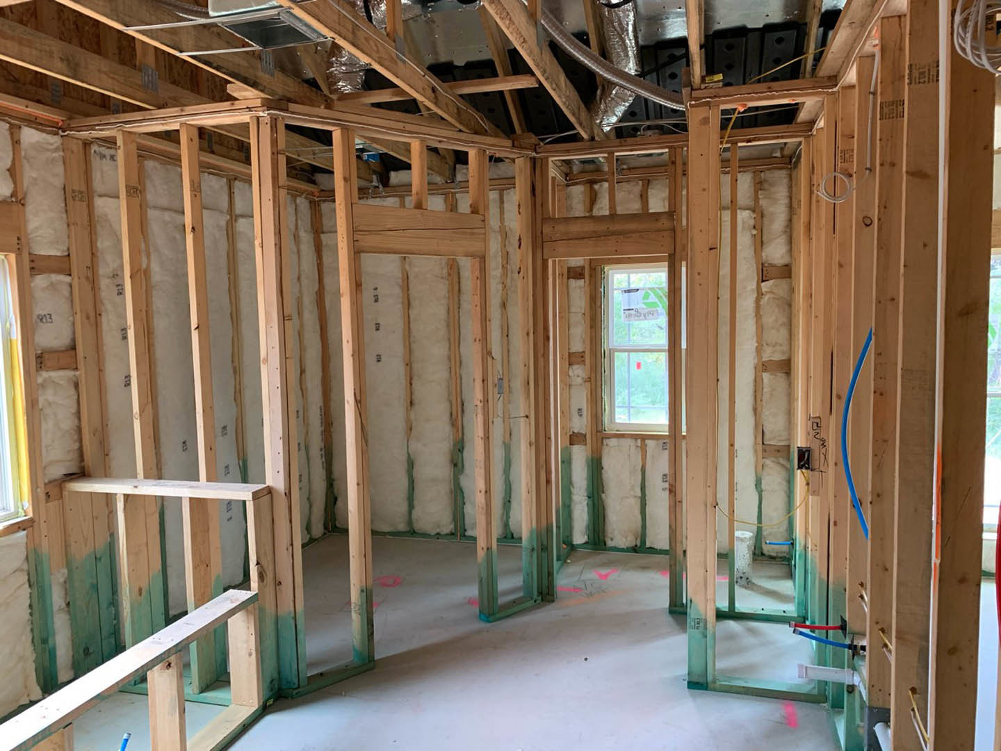 Wood-framed room under construction with exposed beams, partially finished wall, metal ceiling panels, window displaying a sign, blue pencil resting on a wooden beam, and white