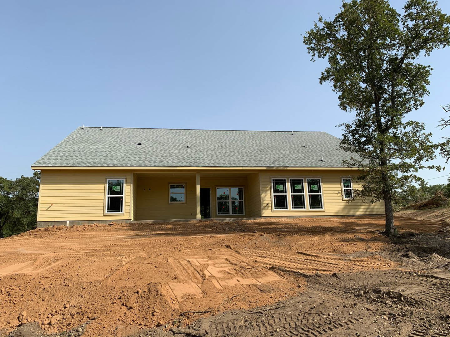 Two-story house under construction with exposed framing, dirt yard marked by tire tracks, large tree in front, covered porch, and window displaying a construction permit sign.