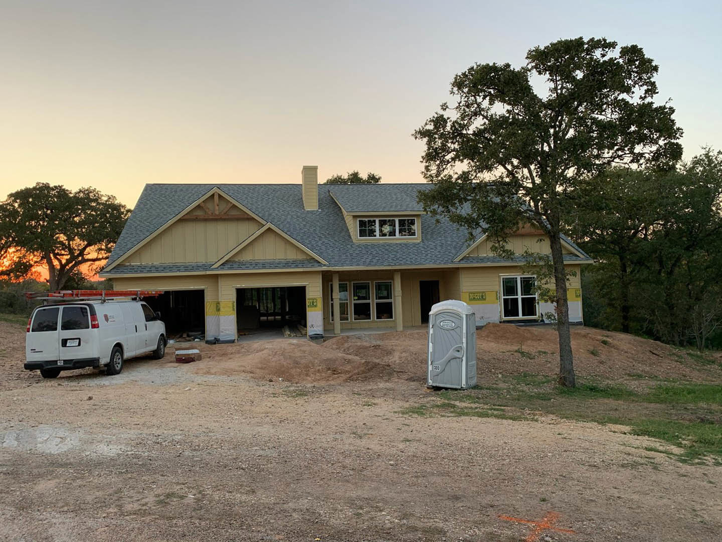 Two-story house with light siding, attached garage, white van with roof ladders parked in driveway, leafy tree beside entrance, portable toilet near construction area, windows