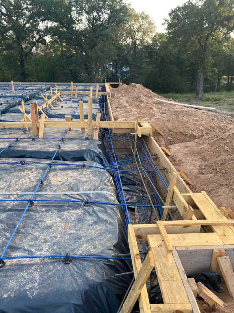 Wooden framing and concrete foundation surrounded by piles of dirt, blue pipes, and black plastic tarp at an outdoor construction site with trees and sky in the background