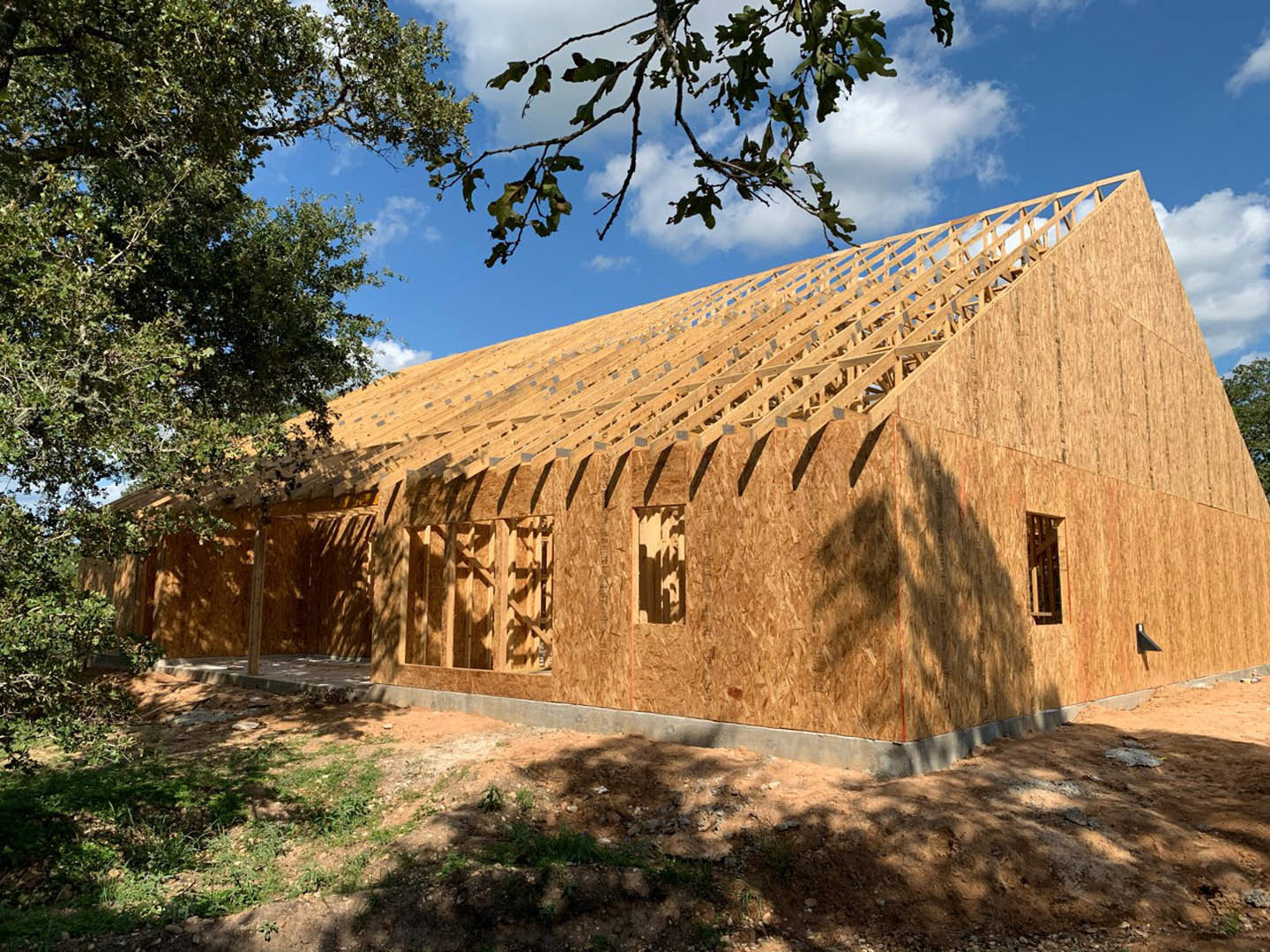 Partially built brick and stone exterior walls surrounded by dirt ground, leafy tree branches, and clear blue sky