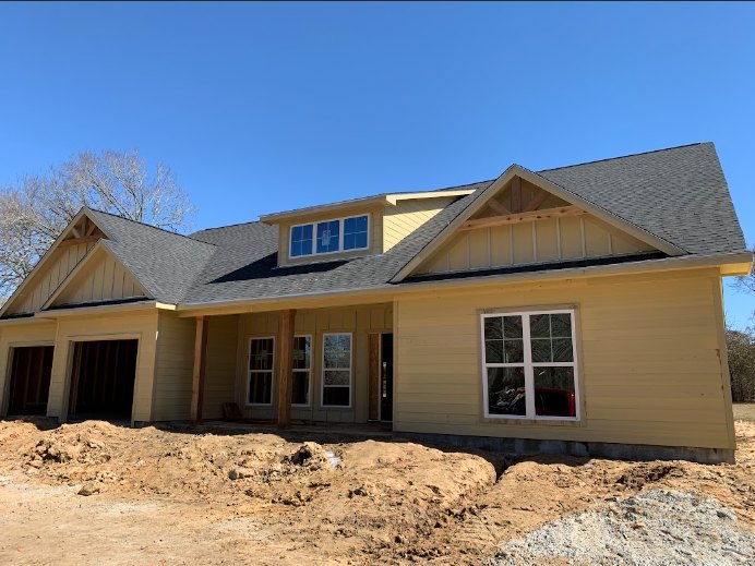 Partially built house with exposed framing, white siding, and dark doorway, surrounded by dirt mounds and trees under a clear blue sky