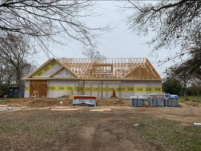 Wood-framed house under construction with exposed beams, pallets stacked near foundation, dirt road leading to site, scattered building materials, trees and open land in background