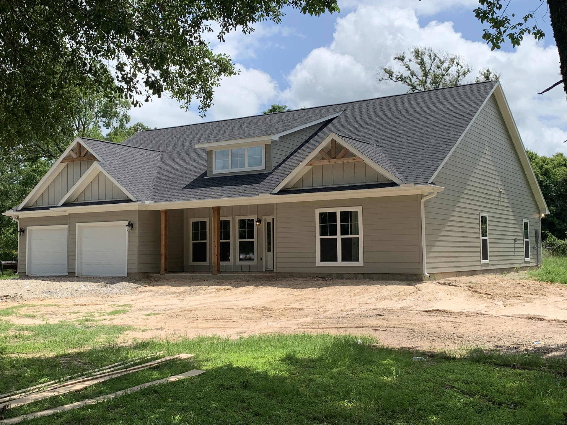 Two-story house under construction with white-framed windows, white garage door, covered porch, and wooden planks scattered on grassy yard; tree partially visible beside sunlit