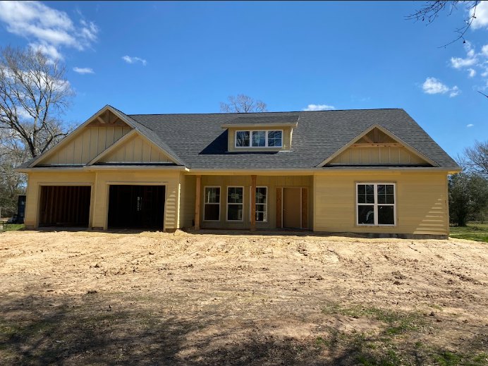 Two-story house under construction with white-framed windows, attached garage, light siding, dirt lot in foreground, and trees in background under clear blue sky