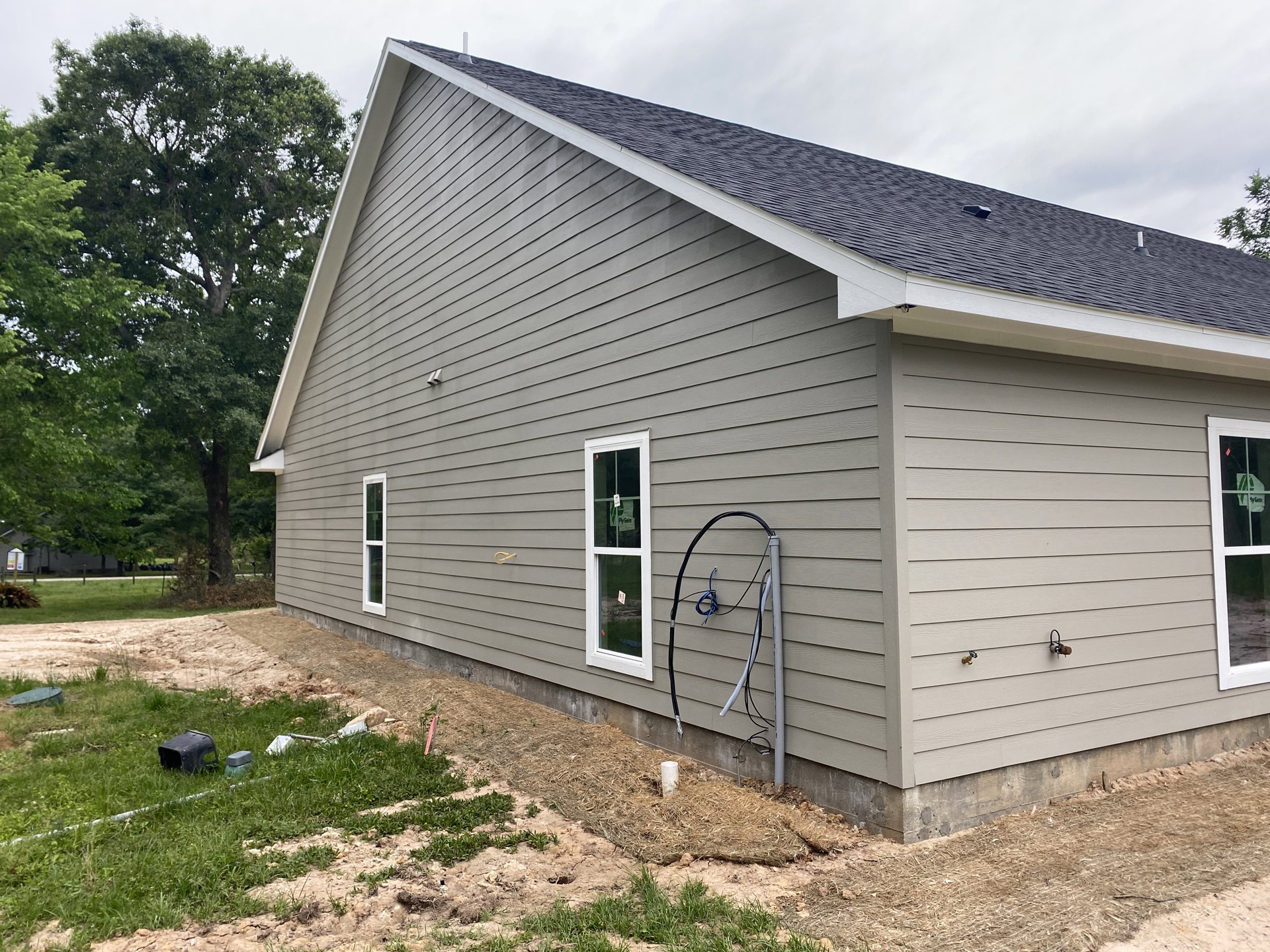 White siding house with garden hose attached to exterior wall, window displaying a sign, electrical wires extending from metal pole, black object resting on grassy lawn, tree