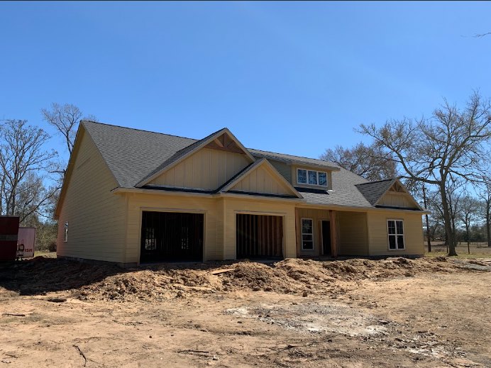 Partially built house with exposed framing, attached garages, dirt piles in front, person standing in doorway, barred window, trees and sky in background
