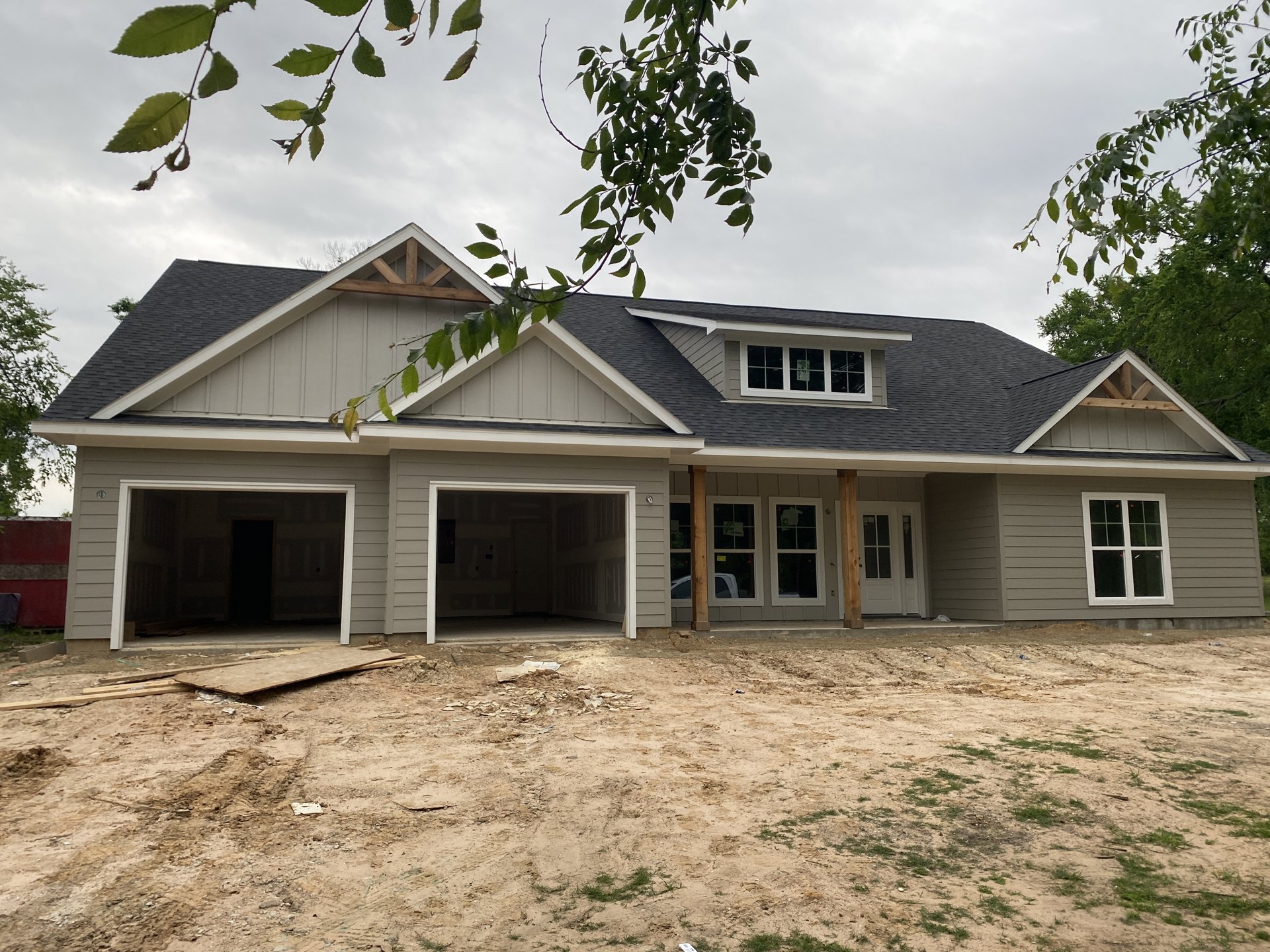 Partially built house with white-framed windows, garage door, green flag in window, surrounded by dirt field and leafy tree branches