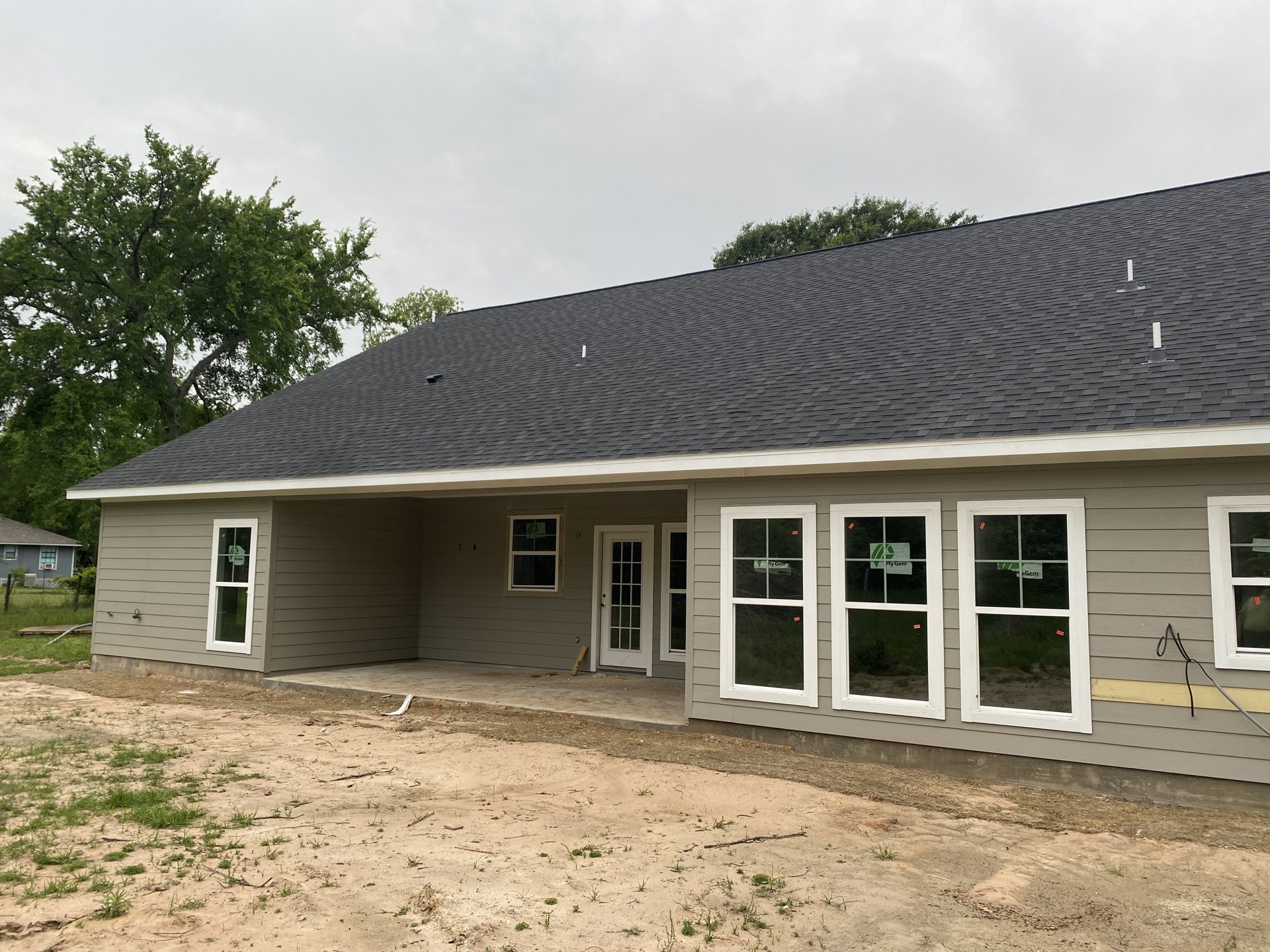 Partially built house with exposed roof and glass-paneled door, dirt road and bare ground in foreground, window displaying red tag, cloudy sky overhead, trees in background.
