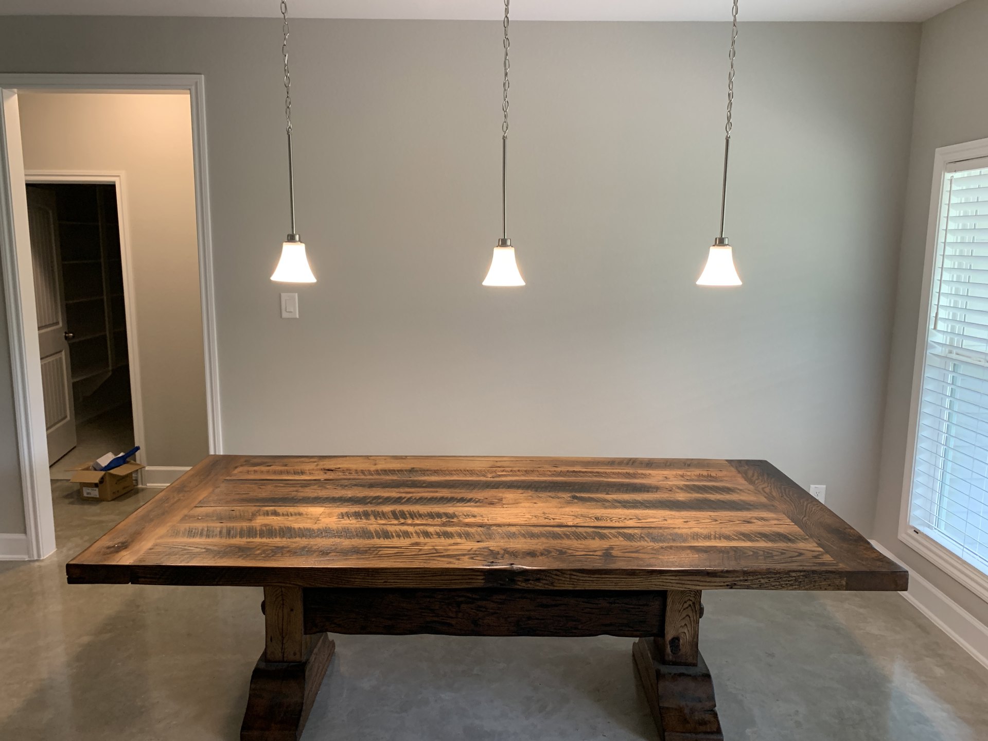 Wooden dining table with angled legs beneath three pendant lights featuring bell-shaped shades, hardwood flooring, window with blinds, open door leading to adjacent room, storage
