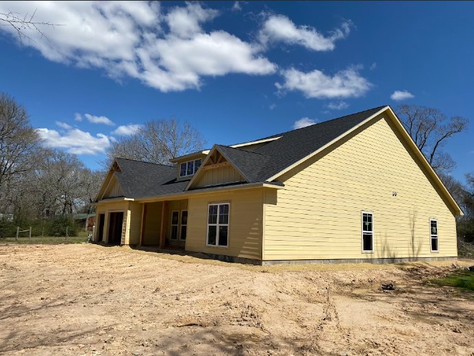 Yellow house with black roof surrounded by dirt patch and mature trees, exterior windows visible under cloudy sky