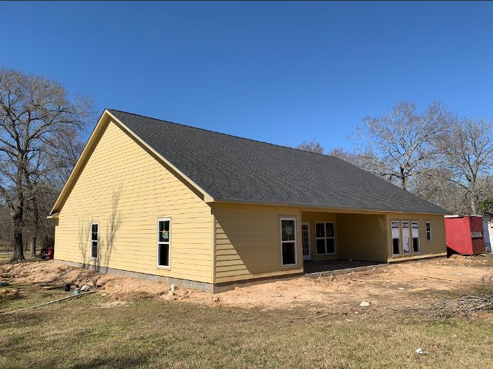 Yellow house with unfinished siding, exposed windows, and construction materials in front; tree nearby and red and black container on site