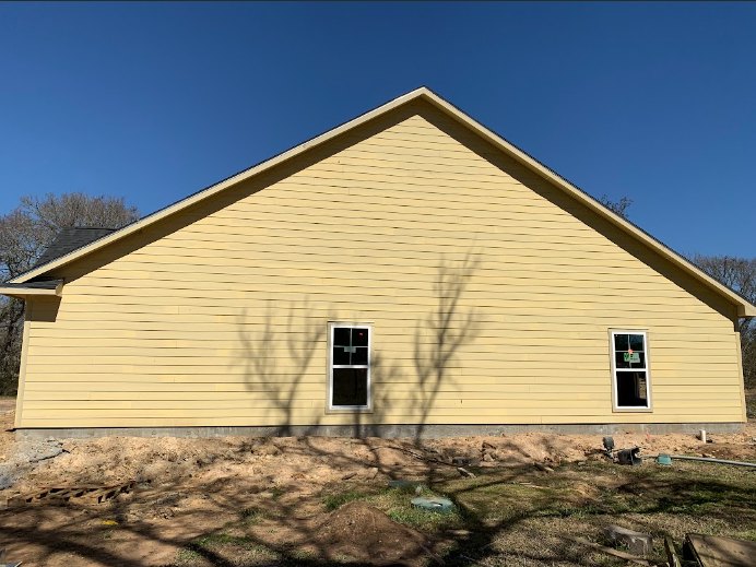 Yellow house with unfinished siding, visible window, pitched roof, and construction materials scattered outdoors