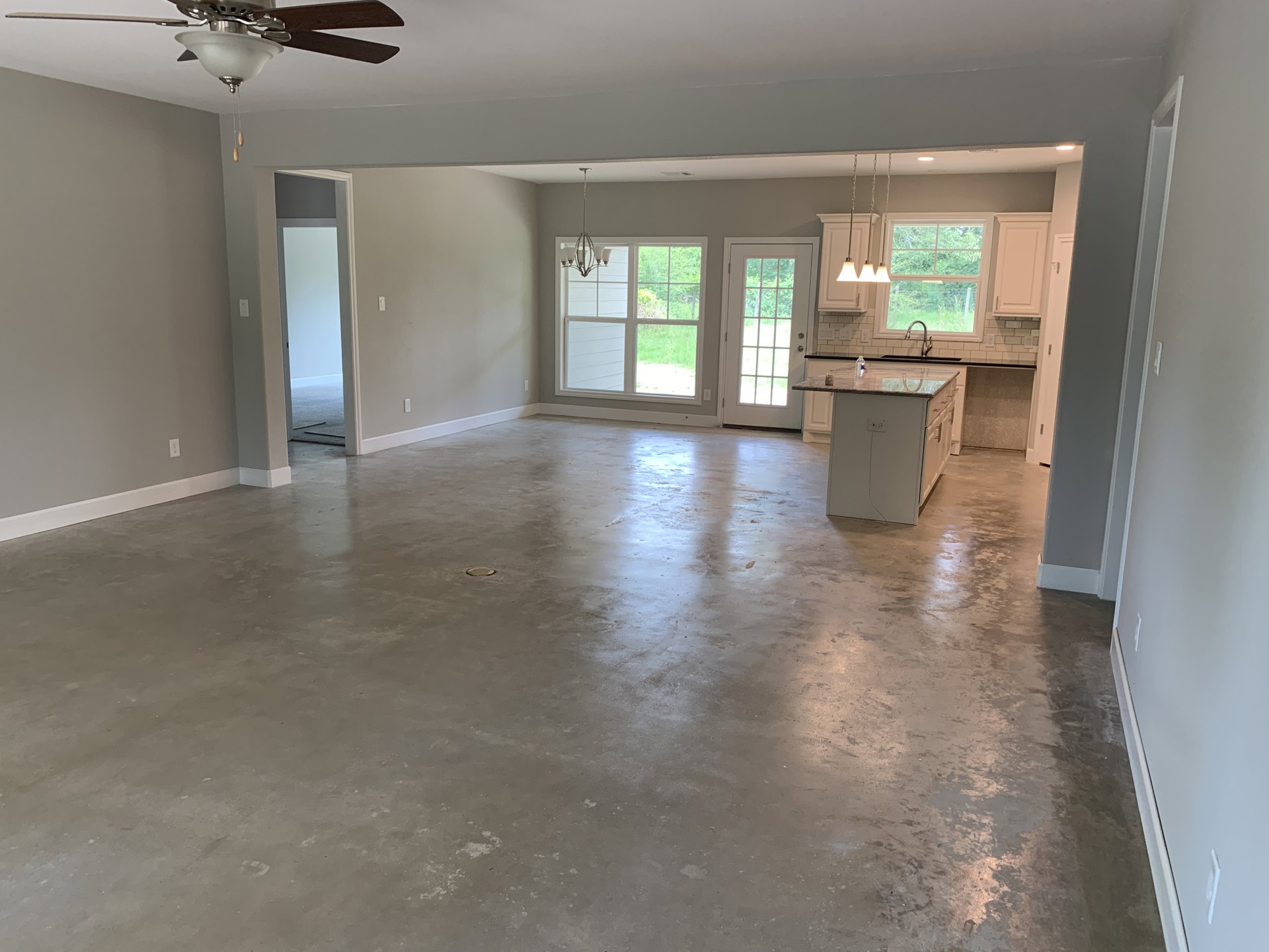 Open-concept living area with concrete flooring, ceiling fan with light fixture, kitchen island holding a water bottle, white plaster walls, glass-paneled door, and window with