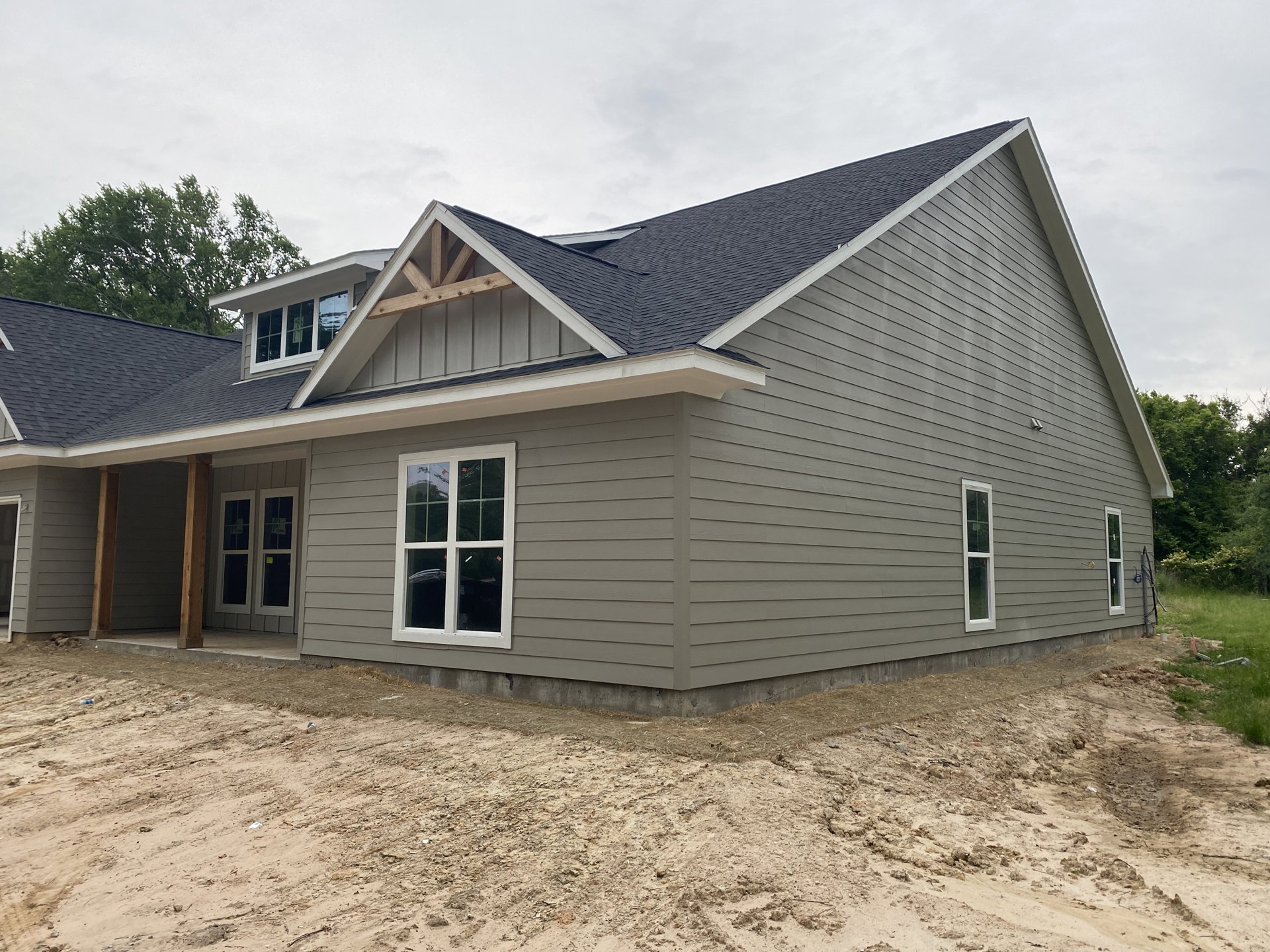 Two-story house under construction with gray siding, white-framed windows featuring yellow stickers, dark shingle roof, dirt lot in foreground, and mature green trees surrounding