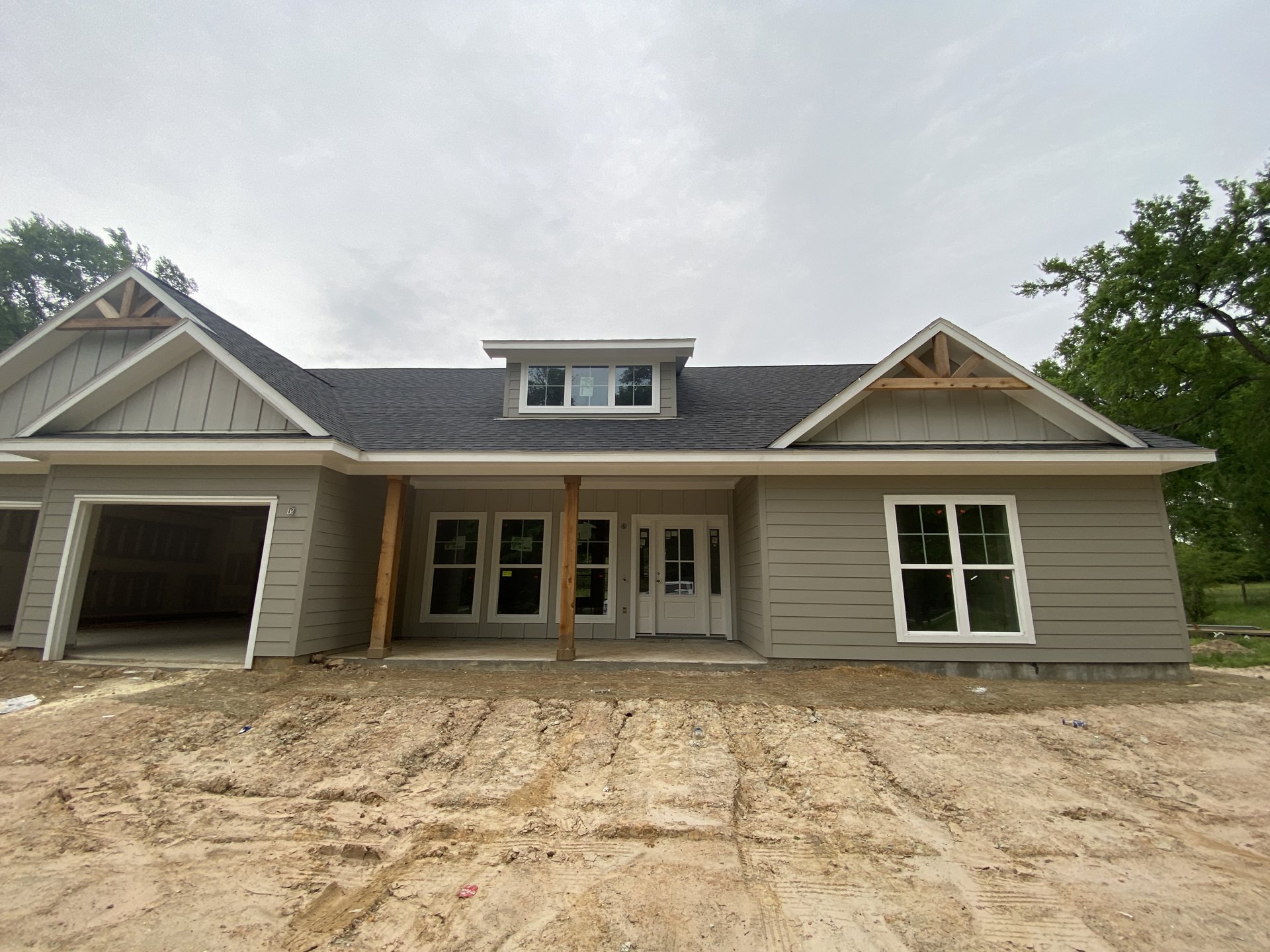 Two-story white house under construction with exposed dirt yard, wooden posts, white-trimmed windows, covered porch, and surrounding trees