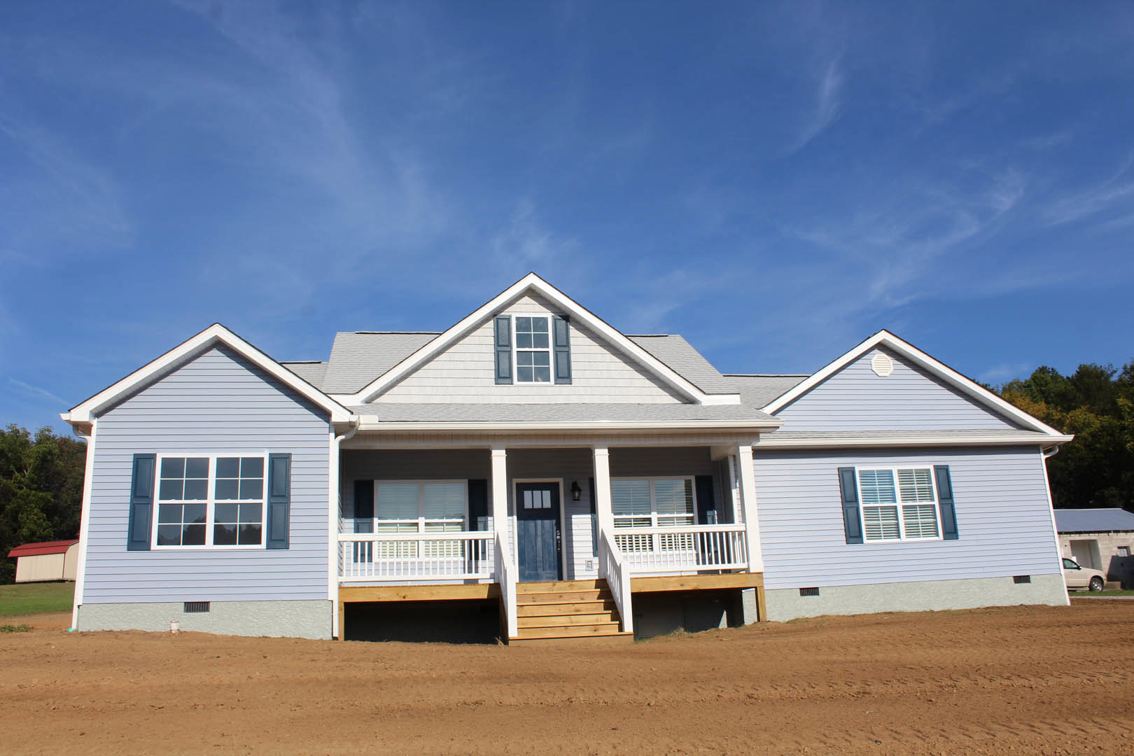 Two-story house with light siding, covered porch, wooden stairs, dirt yard, and blue sky overhead