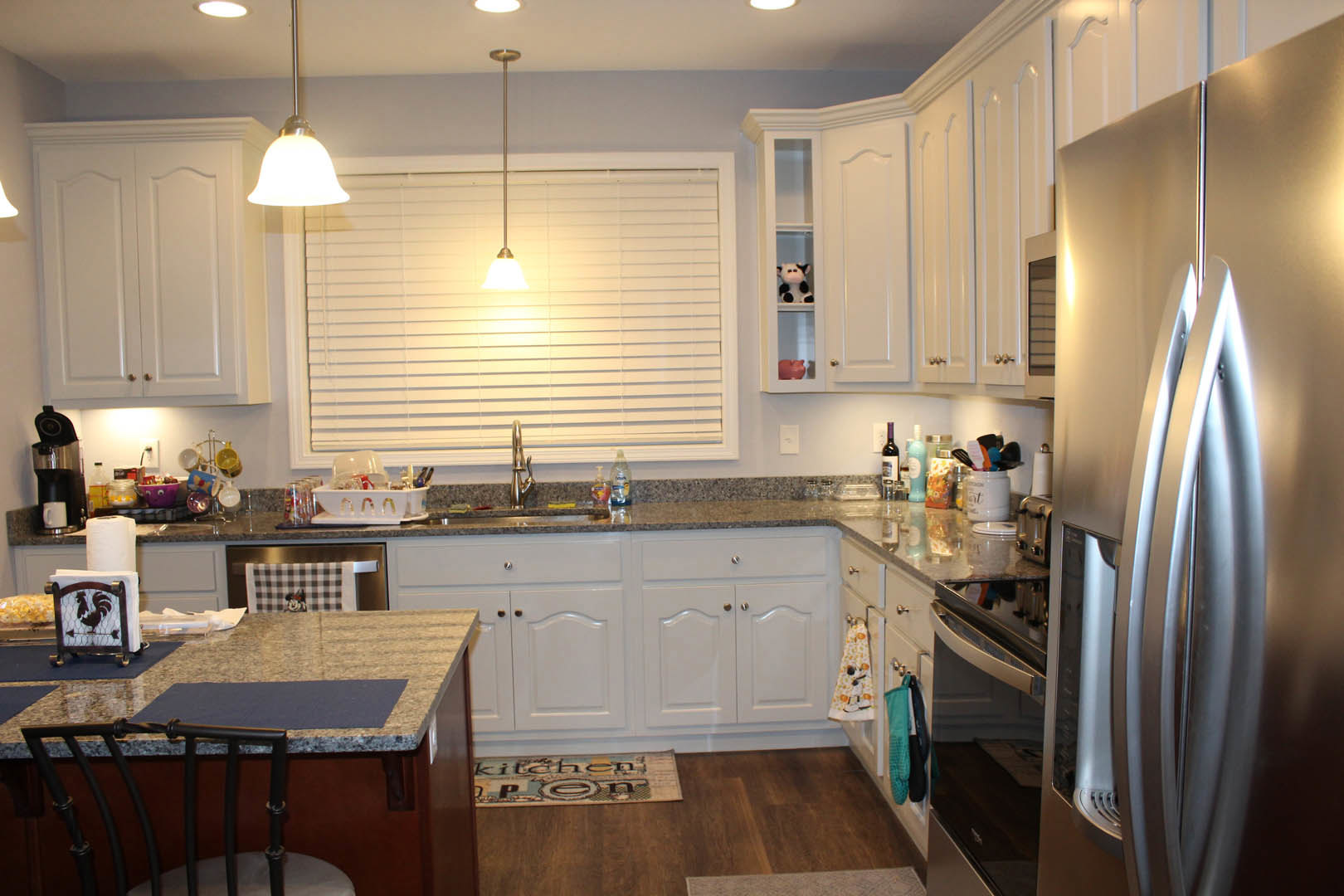 White kitchen cabinets with polished granite countertops, stainless steel refrigerator, ceiling light fixture, wooden chair, sink, and a white jar holding a toothbrush on the
