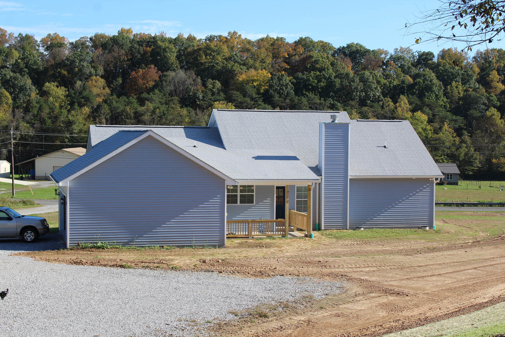 Two-story cottage-style home with gray siding, covered porch featuring wooden and metal railings, gravel driveway in front, parked car partially visible, surrounded by mature trees
