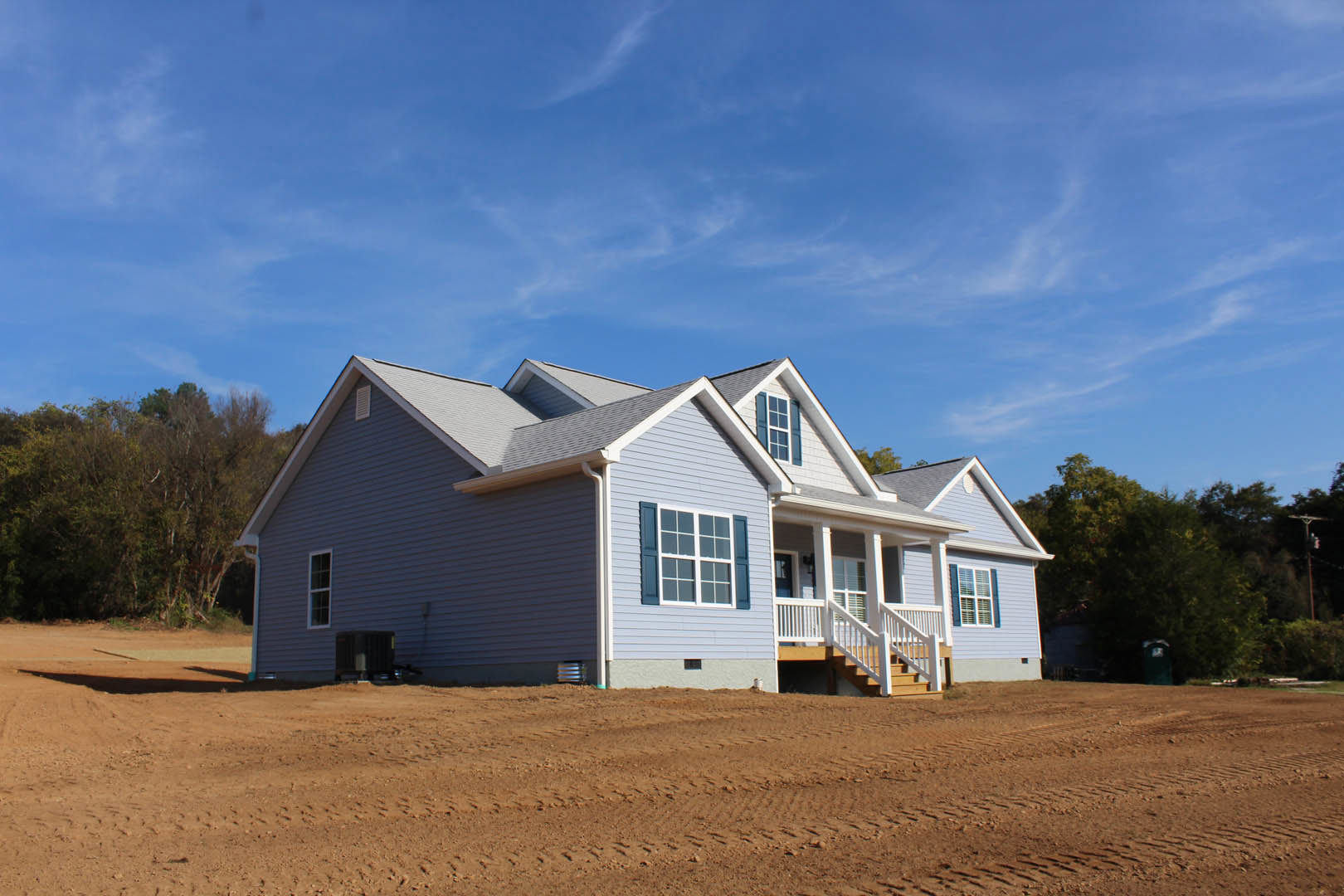 Single-story house with blue roof and white siding, large windows, surrounded by a dirt field with tire tracks, trees and blue sky in the background