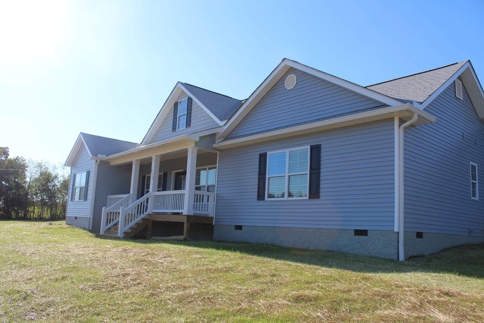 Two-story house with gray siding, white porch railing, large windows, and manicured green lawn bordered by trees.