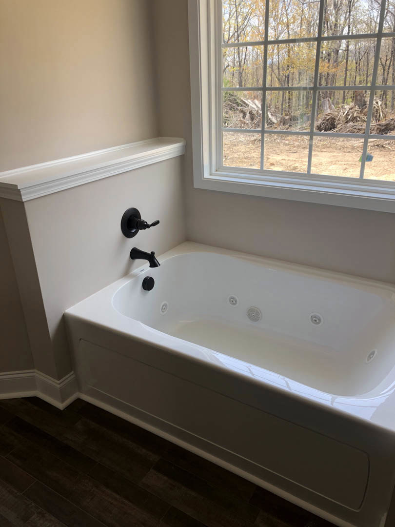 Freestanding white bathtub with matte black faucet and handles, positioned near a window overlooking trees, set against light walls in a modern bathroom.