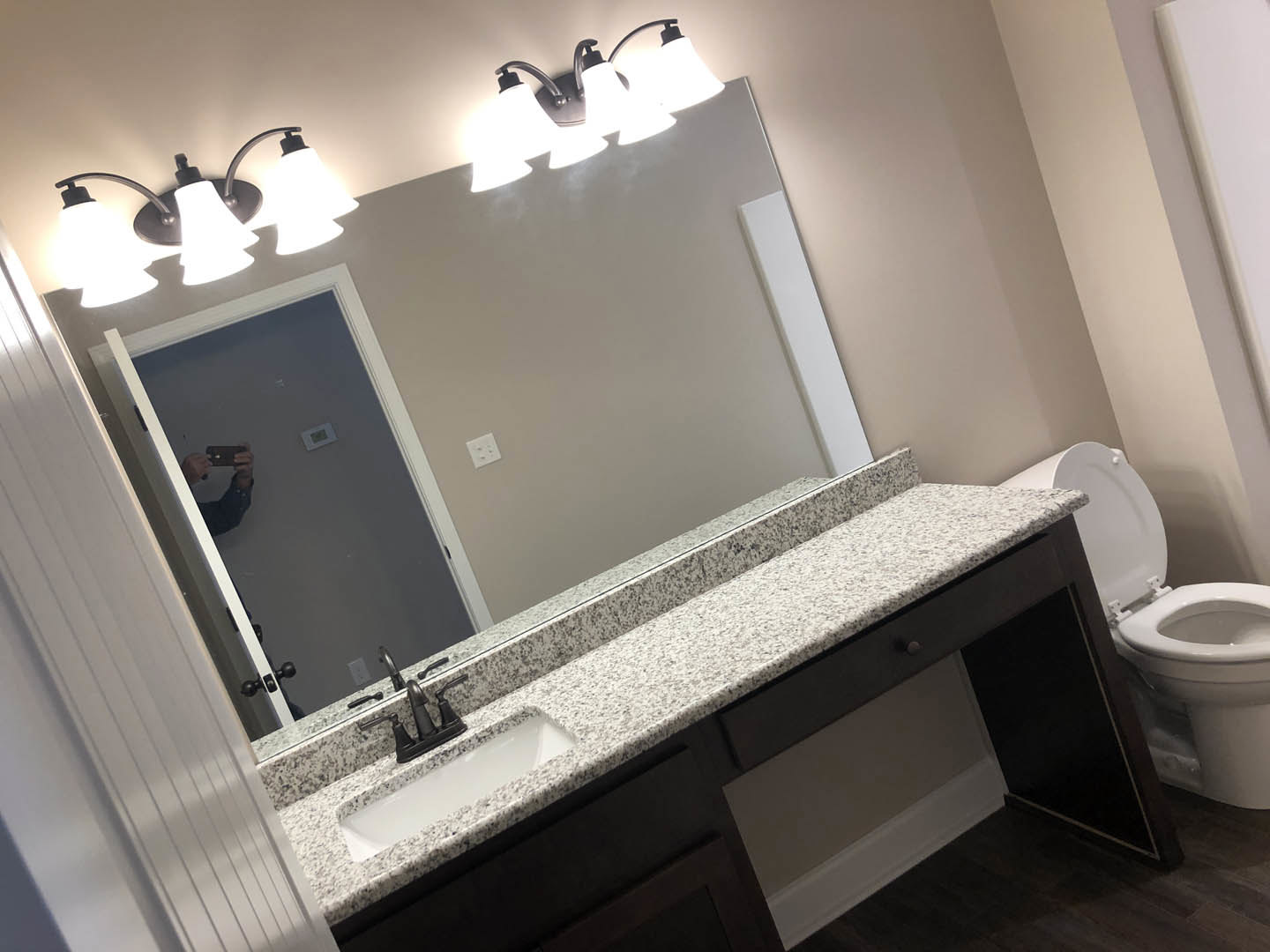 White tile bathroom with rectangular mirror above a modern sink and chrome faucet, toilet with lid up, and overhead light fixture; person visible taking a selfie in the mirror.