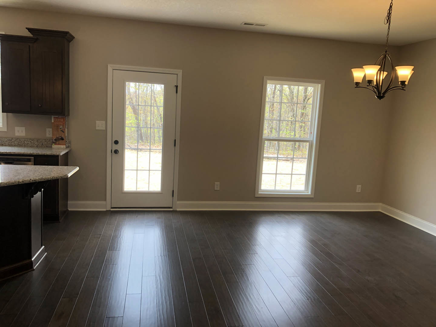 Open kitchen with dark wood flooring, white cabinetry, stone countertops, large window overlooking trees, and modern chandelier hanging above island
