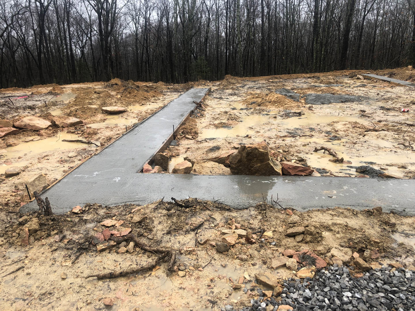 Concrete walkway set in bare dirt, bordered by scattered rocks and soil, with a backdrop of mature trees and open sky.